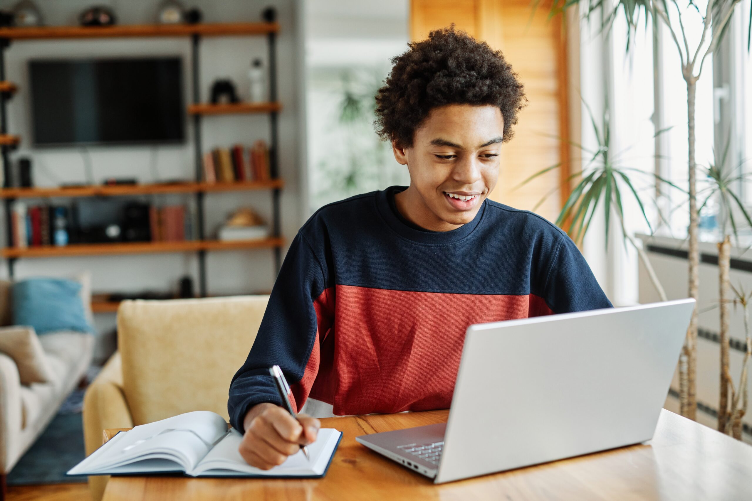 teenage boy smiling at an open laptop while he writes in a notebook