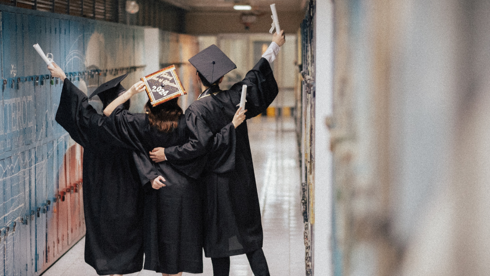 three students clad in graduation caps and gowns posing in a high school hallway