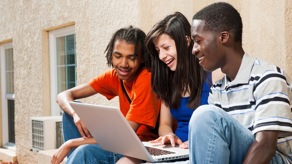three students sitting outside and smiling at a laptop together