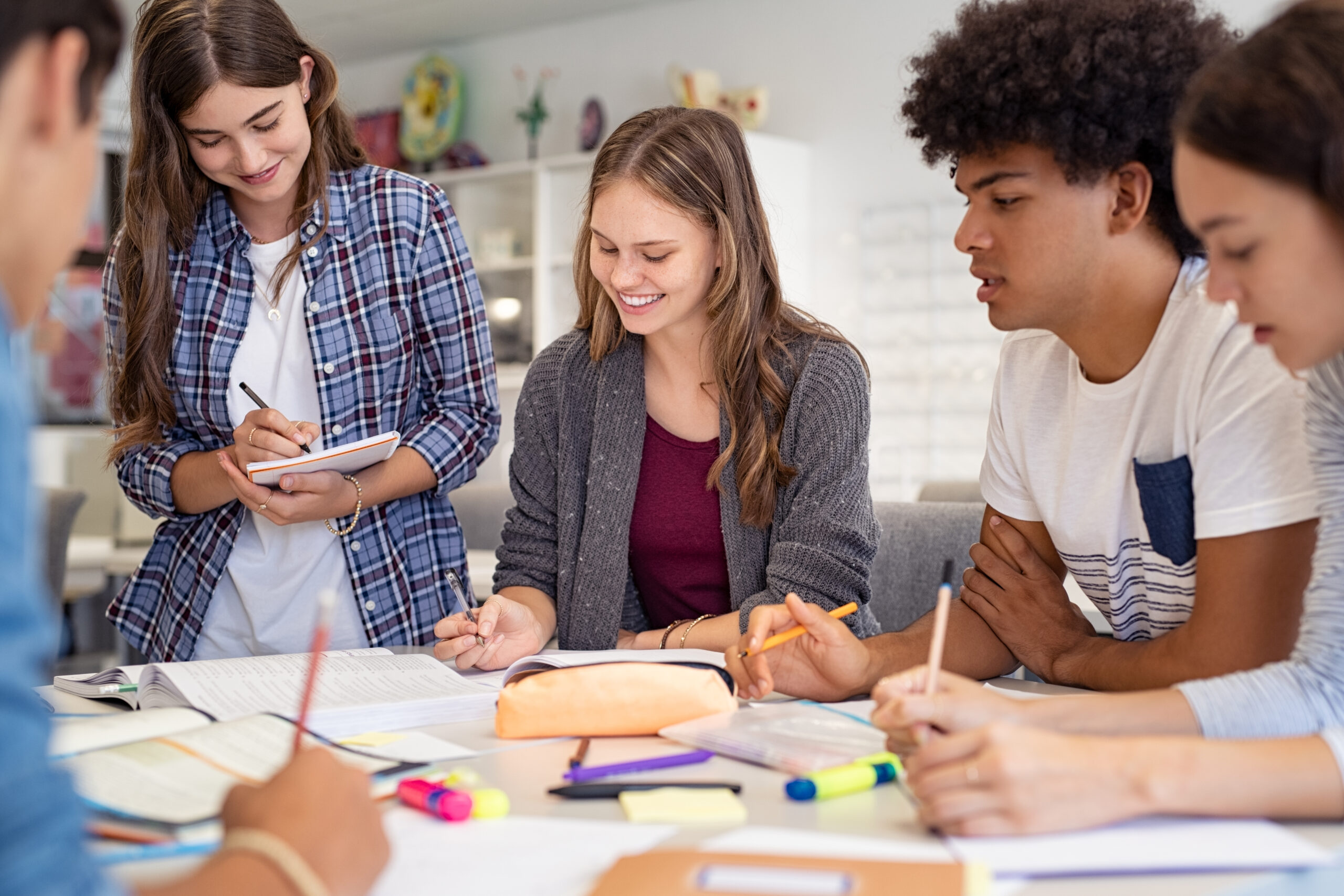 Five high school students gathered around a table, each with paper and pens. They seem to be working on a group project.