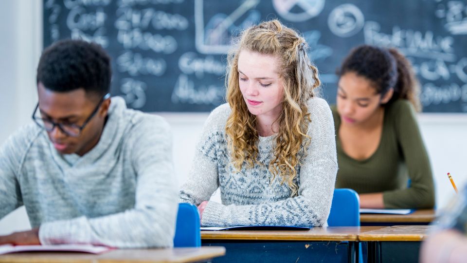 three students in a classroom, looking down as they write on paper at their desks