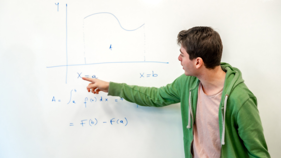 a student in a green hooded sweatshirt pointing at a math equation on a whiteboard