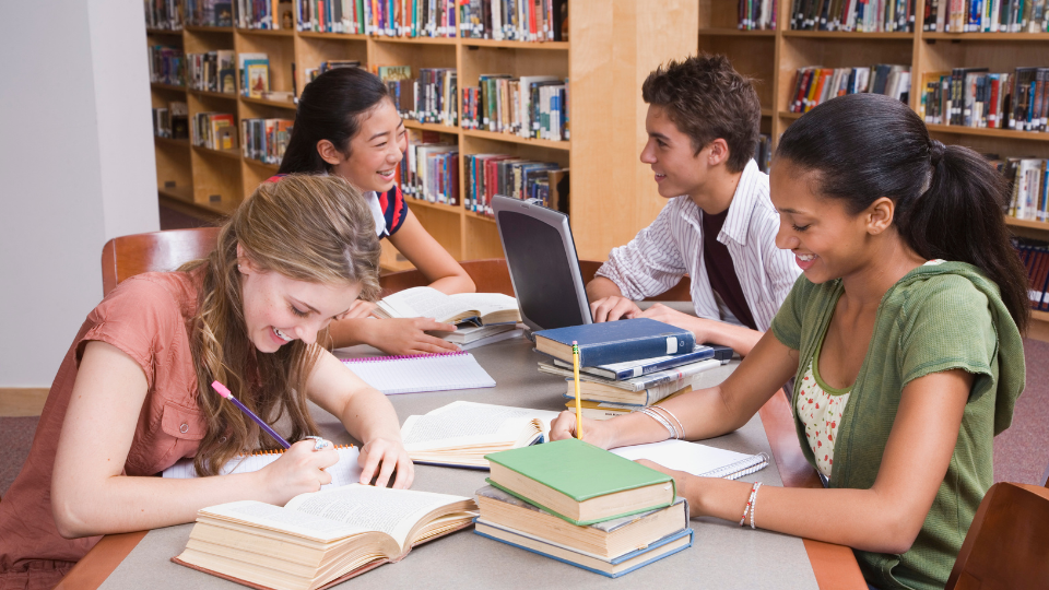 group of four high school students around a table, covered in books and laptops, in a library