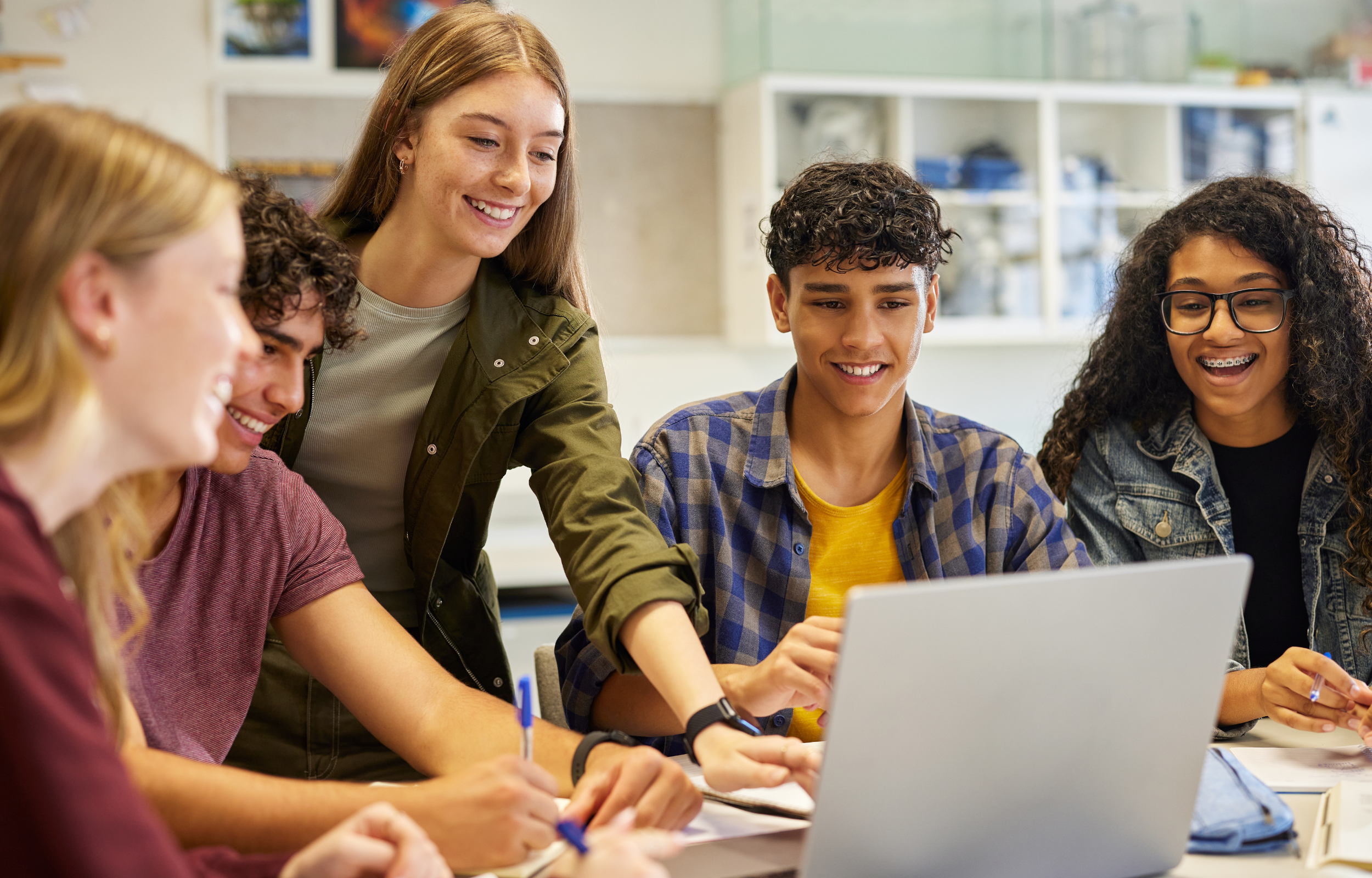 students gathered around a table, smiling together at a laptop